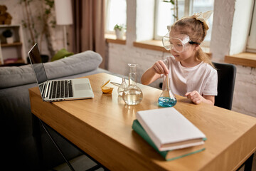 Girl, elementary school pupil wearing safety goggles, conducting home experiment with blue liquid, learning science basics through interactive online class. Concept of education, remote studying