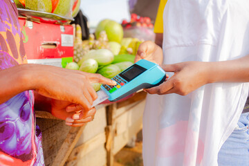 Person paying for fruits with a credit card while african fruit seller woman holding an electronic...