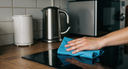 Hand wiping black induction cooktop with blue cloth. Cleaning kitchen appliance surface. Housekeeping concept for kitchen maintenance, appliance care and daily cleaning routine