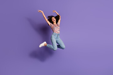 Joyful young woman with beautiful curls jumping playfully against purple background, showcasing vibrant fashion