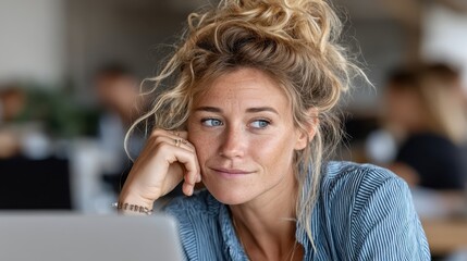 Fototapeta premium A woman with a messy bun gazes thoughtfully off-camera, resting her chin on her fist while her laptop sits before her, embodying creativity and contemplation in a cozy home setting.