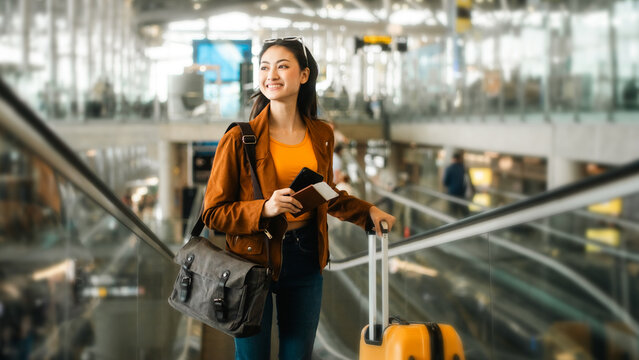 happy young asian woman traveler holding boarding pass ticket, passport and smartphone at the airport terminal with her luggage and smiling at camera, cheerful tourist female having holiday trip.