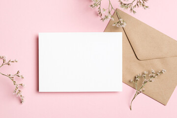 Horizontal white blank card mockup with craft envelope and delicate flowers on soft pink background, ideal for wedding invitation, flat lay with copy space