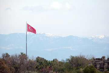 Turkish state flag waving on flagpole before rural landscape with high mountains with snow on the tops at far in light haze