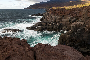Waves crash against the rocks against the background of a volcano. Iturup Island, Kuril Islands