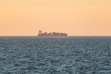 A large cargo ship with containers sails on the blue sea under a warm orange sky at sunset.