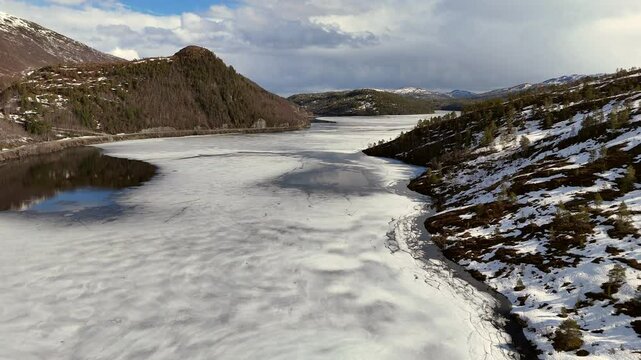 Aerial view of the frozen lake Vasslivatnet surrounded by snowy hills and mountains under cloudy sky