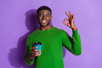 Young stylish man in green pullover holding coffee cup and showing okay gesture against purple...