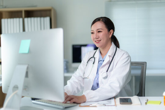 Asian female doctor wearing lab coat and stethoscope working on desktop computer in medical office