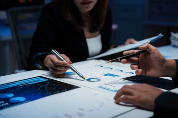Business colleagues examining financial reports, pointing at charts and graphs with pens, working late in office