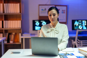 Asian businesswoman concentrating on working late at night using laptop in office with computer screens showing charts and graphs