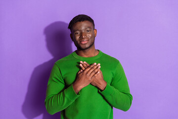 Young man wearing green sweatshirt expressing gratitude with hands over heart in front of a vivid purple background
