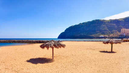 Tranquil beach scene on Madeira, Portugal, with palm umbrellas casting a shadow on golden sand under a clear blue sky. Island vacation concept.