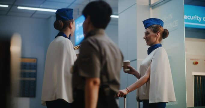Airline Pilot and Two Beautiful Flight Attendants in Uniforms with Luggage Talking, Drinking Coffee, Waiting for Commercial Flight. Flight Crew in Crowded International Airport Terminal. Slow Motion.