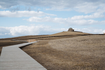 Obraz premium A beautifully crafted wooden walkway gracefully traverses a vast expanse of dry, golden grassy field, inviting those who wander to explore its serene landscape of natural beauty