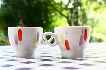 On a table with a checkered cloth there are two cups with original decorating. Garden plants serve as a green background.