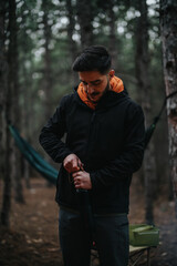 Fototapeta premium Image of a man opening a bottle while enjoying a picnic outdoors in a forest setting during wintertime, featuring equipment like a hammock and a cooler in the background.