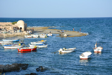 Fototapeta premium Fishermen village in Tunisia. Serene coastal scene with several fishing boats anchored in the clear blue water. A rocky shoreline extends into the sea, with people fishing at the water's edge.