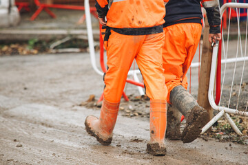 Two construction workers dressed in bright orange gear walk at muddy worksite, surrounded by barriers and construction equipment