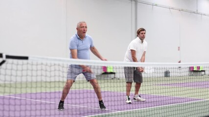 Portrait of sporty adult man playing doubles pickleball with experienced aged partner on indoor court, ready to hit ball. Sport and active lifestyle concept