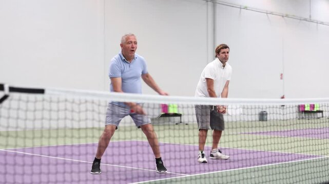 Portrait of sporty adult man playing doubles pickleball with experienced aged partner on indoor court, ready to hit ball. Sport and active lifestyle concept