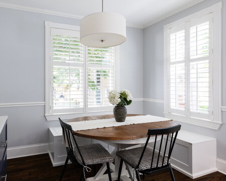 A cozy dining room with blue walls and white plantation shutters, black chairs and white bench seating around a wood table, and a white light fixture hanging above.