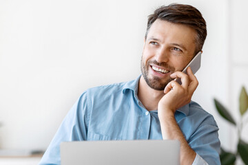 Handsome Male Entrepreneur Having Phone Conversation And Drinking Coffee At Workplace, Sitting At Desk With Laptop In Office, Having Break At Work, Enjoying Mobile Communication, Copy Space