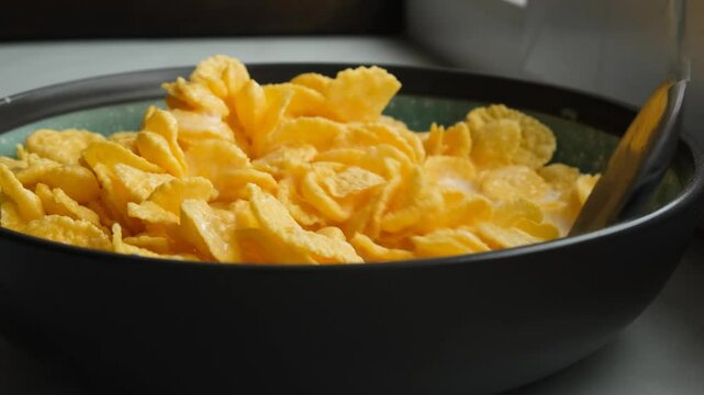 A metal tablespoon stirs yellow cornflakes into milk in a bowl. Mix cornflakes and fresh milk thoroughly for a nutritious low-calorie breakfast. Vegetarianism and diet.