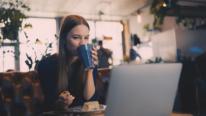 Young adult woman reading news on laptop while having breakfast in cozy cafe