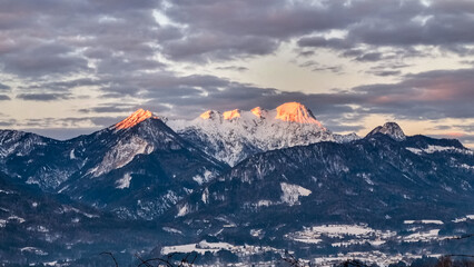 Majestic view of Mittagskogel in Karawanks mountain range, Carinthia, Austria, illuminated by the warm glow of sunset. Snow-covered peaks rise above a serene valley dotted with alpine villages