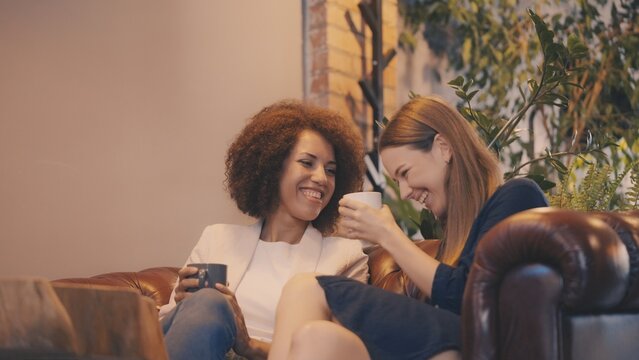 Two female friends sharing sincere laughter and enjoying coffee in cozy coffeehouse