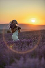 Blonde woman poses in lavender field at sunset. Happy woman in white dress holds lavender bouquet. Aromatherapy concept, lavender oil, photo session in lavender