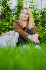 Fototapeta premium Young woman sitting on a grassy lawn with a backpack, brown jacket, black pants, background is garden or park with bushes. the overall mood of the image is peaceful and relaxed.
