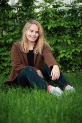 Fototapeta premium Young woman sitting on a grassy lawn with a backpack, brown jacket, black pants, background is garden or park with bushes. the overall mood of the image is peaceful and relaxed.
