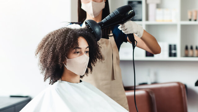 Social distancing concept. Young hairdresser in protective mask making stylish hairdo for african american customer, empty space