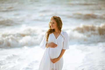 Happy pregnant woman wearing white dress posing over sea with waves outdoors. Summer vacation...