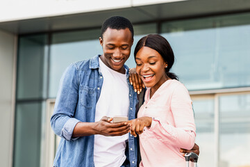 Black travellers couple using smartphone outdoors, standing near airport building, happy african...