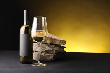 Bottle of wine, wineglass and rocks on black table against dark yellow background, space for text