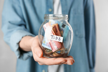 Woman holding tips jar with money on light background, closeup