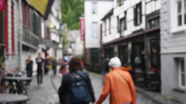 Blurry footage of elderly couple holding hands as they walk down city cobblestone street, sharing intimate moment of togetherness against backdrop of bustling cityscape and ancient architecture,