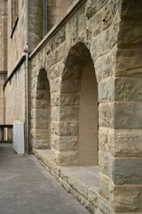 Historic stone building exterior with arched passages, textured sandstone walls, and trimmed bushes in a quiet courtyard. Classic architectural details suitable for heritage, university, and European 