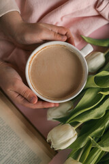 Close-up of a child's hands gently holding a cup of coffee surrounded by white tulips, an open book, and soft pink fabric, creating a calm and cozy atmosphere