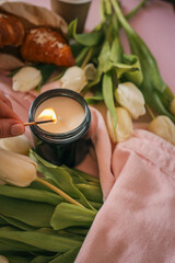 Close-up of a hand lighting a candle with a match. Soft focus on white tulips and a pink jacket in...