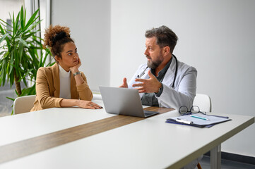 Obraz premium Male doctor with laptop on desk explaining medical procedure to serious female patient in medical clinic