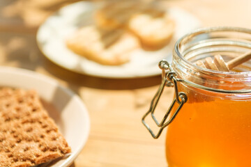 A jar filled with golden honey sits on a wooden table next to crispy crackers and a plate of toasted bread, bathed in warm sunlight, evoking a cozy atmosphere.