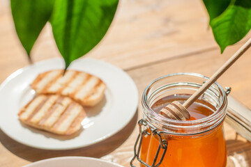 A jar of honey sits with a wooden dipper next to grilled bread slices, illuminated by soft natural light in a cozy kitchen, evoking a warm and inviting atmosphere.