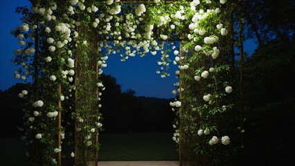 A floral archway in the evening light, adorned with cascades of white blossoms and lush greenery leading to the dark forest.