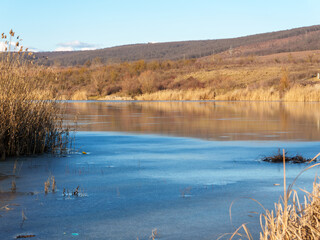 A serene river landscape features a thin layer of ice covering the water. Tall golden reeds line the riverbank under a bright blue sky, with distant hills adding depth to the scene.