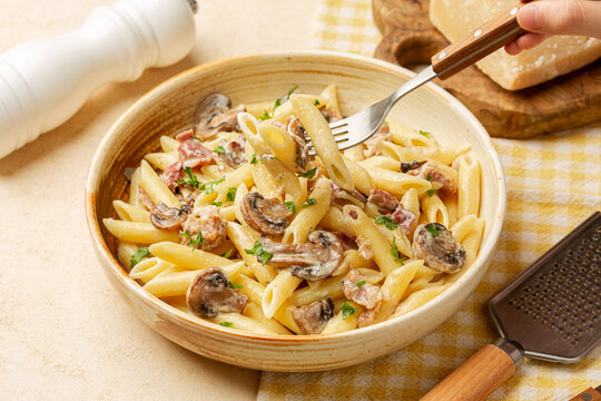 Eating creamy pasta penne with mushrooms and bacon. Hard cheese and grater. Beige table surface.