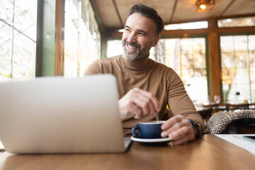 A smiling man stirring his coffee while working on his laptop, reflecting a perfect blend of productivity and enjoyment.